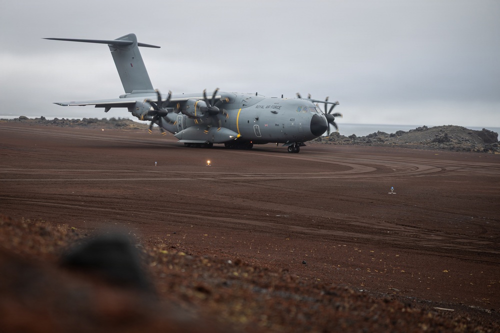 A Royal Air Force Airbus A400M Atlas military transport aircraft is taxiing or stationary on a remote, unpaved or minimally developed airstrip. The terrain is characterized by red-brown volcanic soil and dark lava rock formations, suggesting a remote island location. The overcast sky and rugged landscape indicate an austere or expeditionary operating environment.