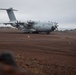 A Royal Air Force Airbus A400M Atlas military transport aircraft is taxiing or stationary on a remote, unpaved or minimally developed airstrip. The terrain is characterized by red-brown volcanic soil and dark lava rock formations, suggesting a remote island location. The overcast sky and rugged landscape indicate an austere or expeditionary operating environment.