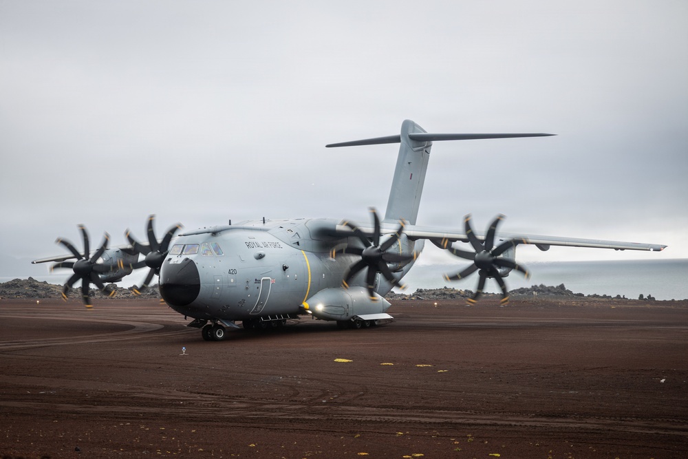 A Royal Air Force Airbus A400M Atlas military transport aircraft, marked with serial number 420, is taxiing on a dark volcanic/dirt runway in an overcast, remote coastal environment. The four turboprop engines with eight-bladed propellers are spinning, indicated by motion blur on the propeller blades. The setting appears to be a remote airstrip, possibly on a volcanic island given the dark terrain and rocky landscape visible in the background.