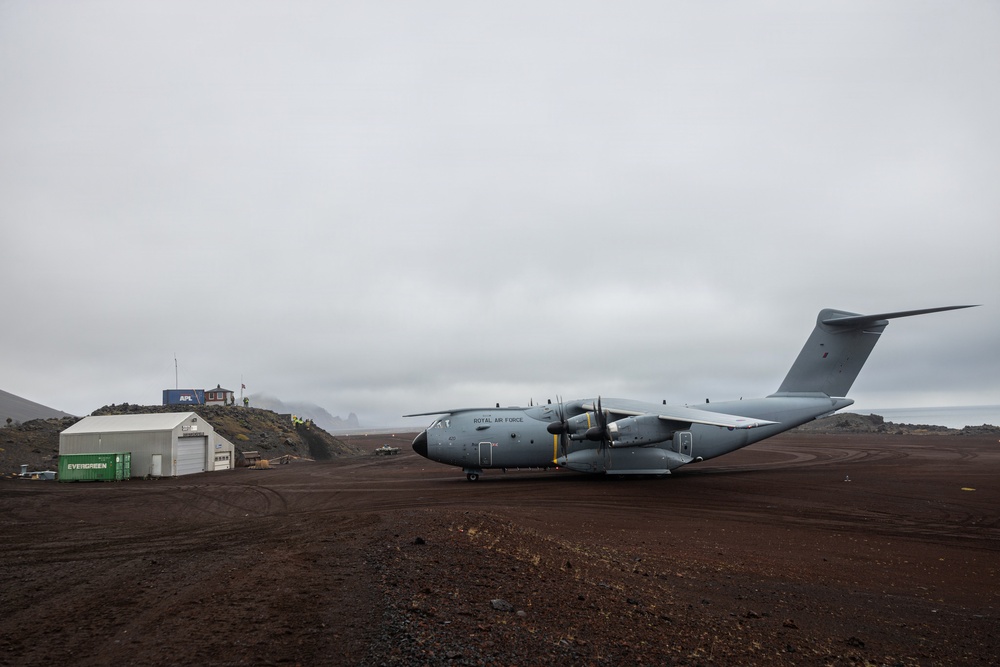 A Royal Air Force Airbus A400M Atlas military transport aircraft is parked on a remote, unpaved airstrip with dark volcanic terrain. The setting appears to be a remote island or polar location, with rugged hills, overcast skies, and minimal infrastructure visible. Several shipping containers and a small hangar building are present in the background.