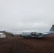 A Royal Air Force Airbus A400M Atlas military transport aircraft is parked on a remote, unpaved airstrip with dark volcanic terrain. The setting appears to be a remote island or polar location, with rugged hills, overcast skies, and minimal infrastructure visible. Several shipping containers and a small hangar building are present in the background.
