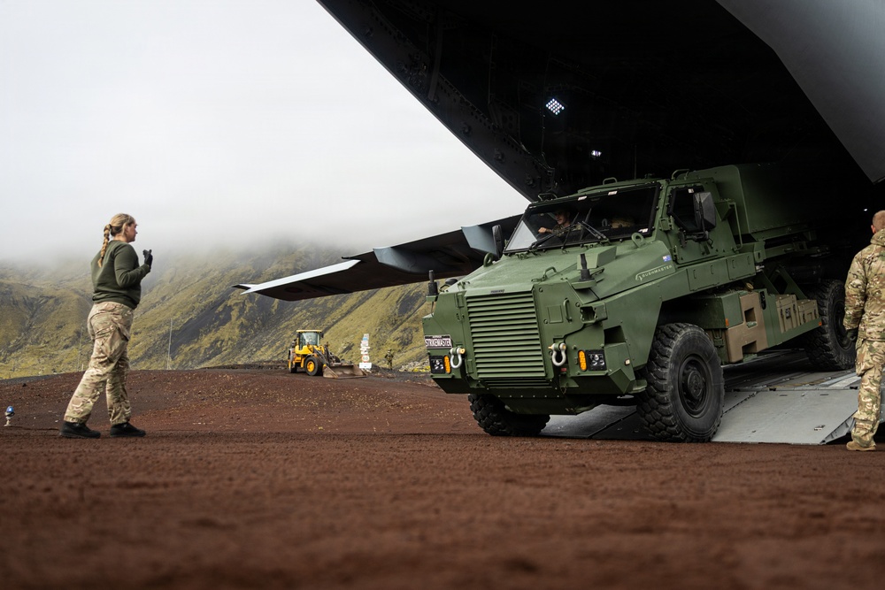 A military armored vehicle is being offloaded from a large transport aircraft via a rear ramp onto a reddish-brown dirt surface. Personnel in military camouflage uniforms are directing the operation, with mountainous terrain visible in the background. A yellow construction loader is also present in the background, suggesting active logistics or base-building operations.