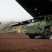 A military armored vehicle is being offloaded from a large transport aircraft via a rear ramp onto a reddish-brown dirt surface. Personnel in military camouflage uniforms are directing the operation, with mountainous terrain visible in the background. A yellow construction loader is also present in the background, suggesting active logistics or base-building operations.