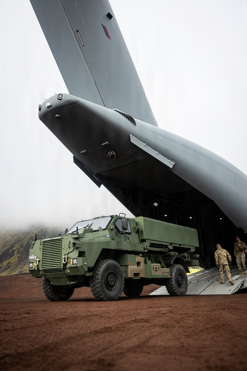A green military protected mobility vehicle, visibly labeled 'Bushmaster' and 'Stingmaster', is being offloaded from the rear ramp of a large military transport aircraft. The aircraft displays a roundel consistent with the Royal Air Force (RAF) on its tail fin. The scene takes place on a red-dirt airstrip in a mountainous, overcast environment.