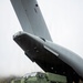 A green military protected mobility vehicle, visibly labeled 'Bushmaster' and 'Stingmaster', is being offloaded from the rear ramp of a large military transport aircraft. The aircraft displays a roundel consistent with the Royal Air Force (RAF) on its tail fin. The scene takes place on a red-dirt airstrip in a mountainous, overcast environment.