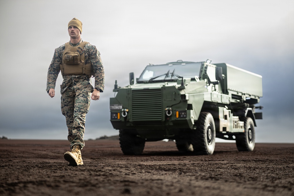A military servicemember in MARPAT digital camouflage uniform and tan tactical vest walks across a dark, muddy terrain toward the camera. Behind him is a green armored military vehicle labeled 'STRIKEMASTER'. The scene is set under an overcast sky, giving the image a dramatic, cinematic quality.