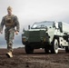 A military servicemember in MARPAT digital camouflage uniform and tan tactical vest walks across a dark, muddy terrain toward the camera. Behind him is a green armored military vehicle labeled 'STRIKEMASTER'. The scene is set under an overcast sky, giving the image a dramatic, cinematic quality.