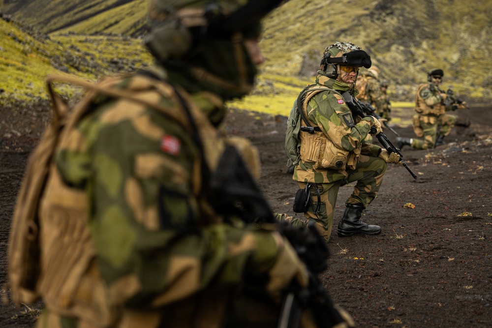 The image shows multiple soldiers in military camouflage uniforms conducting what appears to be a field training exercise or tactical operation in a rugged, volcanic or mountainous terrain. The soldiers are equipped with helmets, tactical vests, and firearms, and appear to be in alert or ready positions. The landscape features dark volcanic soil and green moss-covered hills in the background.