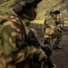The image shows multiple soldiers in military camouflage uniforms conducting what appears to be a field training exercise or tactical operation in a rugged, volcanic or mountainous terrain. The soldiers are equipped with helmets, tactical vests, and firearms, and appear to be in alert or ready positions. The landscape features dark volcanic soil and green moss-covered hills in the background.