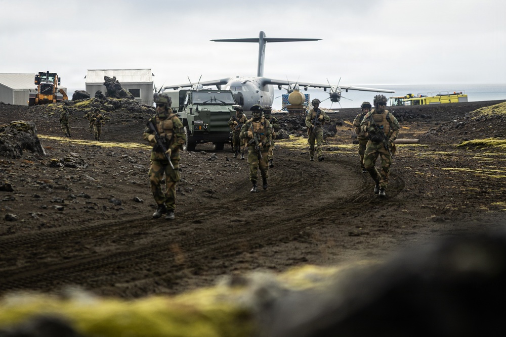 A group of armed soldiers in camouflage uniforms are advancing along a dirt track in a volcanic, lava-field terrain. Behind them, a large military turboprop transport aircraft — consistent with an Airbus A400M Atlas — is parked on what appears to be a remote airstrip. The scene suggests a military exercise or tactical deployment operation in a Nordic or volcanic island environment.