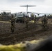 A group of armed soldiers in camouflage uniforms are advancing along a dirt track in a volcanic, lava-field terrain. Behind them, a large military turboprop transport aircraft — consistent with an Airbus A400M Atlas — is parked on what appears to be a remote airstrip. The scene suggests a military exercise or tactical deployment operation in a Nordic or volcanic island environment.