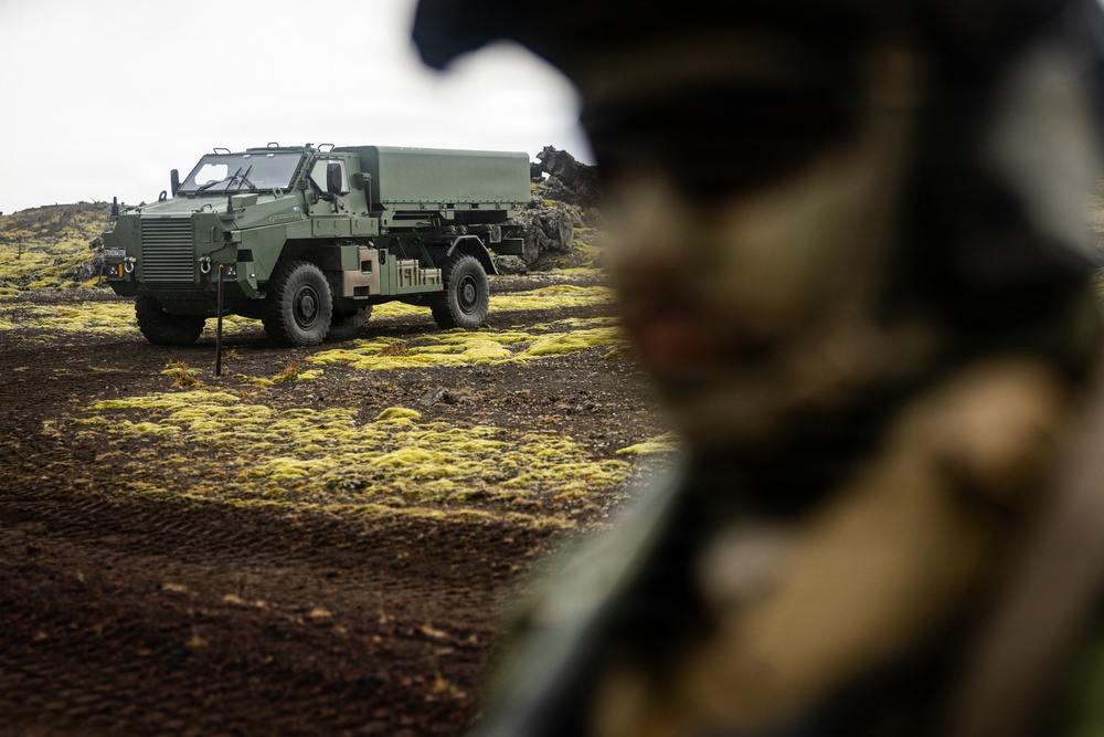 A military armored vehicle, appearing to be an olive/military green protected mobility vehicle, is parked on dark volcanic terrain with patches of yellow-green moss or lichen. In the foreground, a blurred figure wearing military gear and a helmet is partially visible. The setting appears to be a volcanic landscape, consistent with Icelandic terrain.