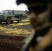 A military armored vehicle, appearing to be an olive/military green protected mobility vehicle, is parked on dark volcanic terrain with patches of yellow-green moss or lichen. In the foreground, a blurred figure wearing military gear and a helmet is partially visible. The setting appears to be a volcanic landscape, consistent with Icelandic terrain.