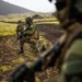 The image shows multiple soldiers in camouflage uniforms conducting what appears to be a tactical patrol or training exercise in a volcanic, moss-covered landscape. At least three soldiers are visible, with the central figure kneeling in a tactical position while holding an assault rifle. The terrain features dark volcanic soil, bright green moss patches, and a dirt track, consistent with Icelandic or similar sub-arctic geography.