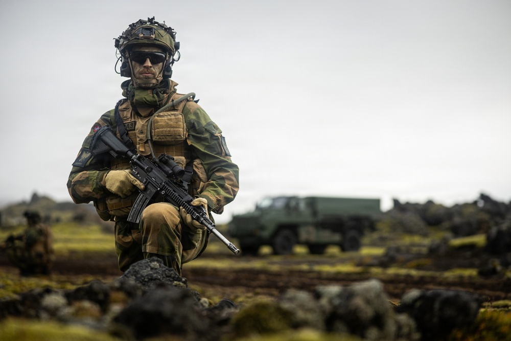 A fully equipped soldier in camouflage uniform kneels on rocky terrain while holding an assault rifle. The setting appears to be a cold, overcast environment with volcanic or rocky ground and sparse vegetation. A military truck is visible in the blurred background.