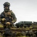 A fully equipped soldier in camouflage uniform kneels on rocky terrain while holding an assault rifle. The setting appears to be a cold, overcast environment with volcanic or rocky ground and sparse vegetation. A military truck is visible in the blurred background.