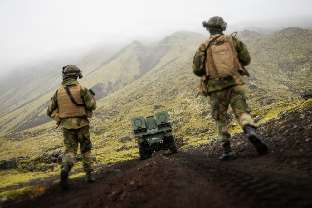 Two soldiers in camouflage uniforms and tactical gear are walking behind a small unmanned ground vehicle (UGV) across a dark volcanic terrain. The landscape features green moss-covered hills partially obscured by low-lying fog or clouds. The scene appears to be a military field exercise or operational patrol in a Nordic or sub-Arctic environment.