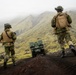 Two soldiers in camouflage uniforms and tactical gear are walking behind a small unmanned ground vehicle (UGV) across a dark volcanic terrain. The landscape features green moss-covered hills partially obscured by low-lying fog or clouds. The scene appears to be a military field exercise or operational patrol in a Nordic or sub-Arctic environment.