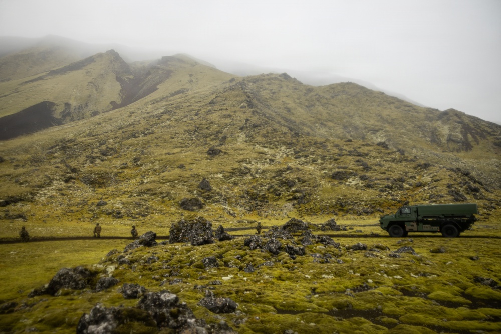 A misty, volcanic highland landscape covered in green moss and scattered lava rocks, with a military-style green truck parked along a road in the middle distance. Several small human figures are visible near the road, suggesting a military or expedition operation. The fog-shrouded mountains in the background create a dramatic, remote atmosphere.