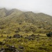 A misty, volcanic highland landscape covered in green moss and scattered lava rocks, with a military-style green truck parked along a road in the middle distance. Several small human figures are visible near the road, suggesting a military or expedition operation. The fog-shrouded mountains in the background create a dramatic, remote atmosphere.