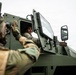 Two military personnel are interacting near a Bushmaster protected mobility vehicle. One soldier is seated inside the vehicle's cab with the armored door open, while another in the foreground gestures toward them. The scene appears to depict a coordination or communication moment during a military exercise or operation.
