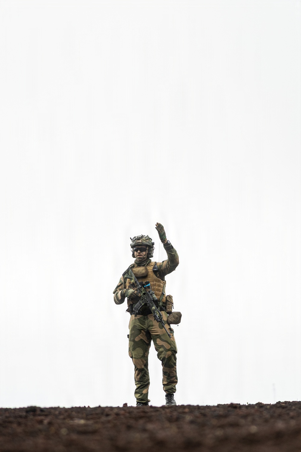 A fully equipped soldier in woodland camouflage stands on an elevated dirt ridge against an overcast white sky. The soldier is raising one hand in a signaling gesture while holding an assault rifle with the other hand. The image is taken from a low angle, giving the figure a prominent silhouette against the pale background.