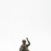 A fully equipped soldier in woodland camouflage stands on an elevated dirt ridge against an overcast white sky. The soldier is raising one hand in a signaling gesture while holding an assault rifle with the other hand. The image is taken from a low angle, giving the figure a prominent silhouette against the pale background.