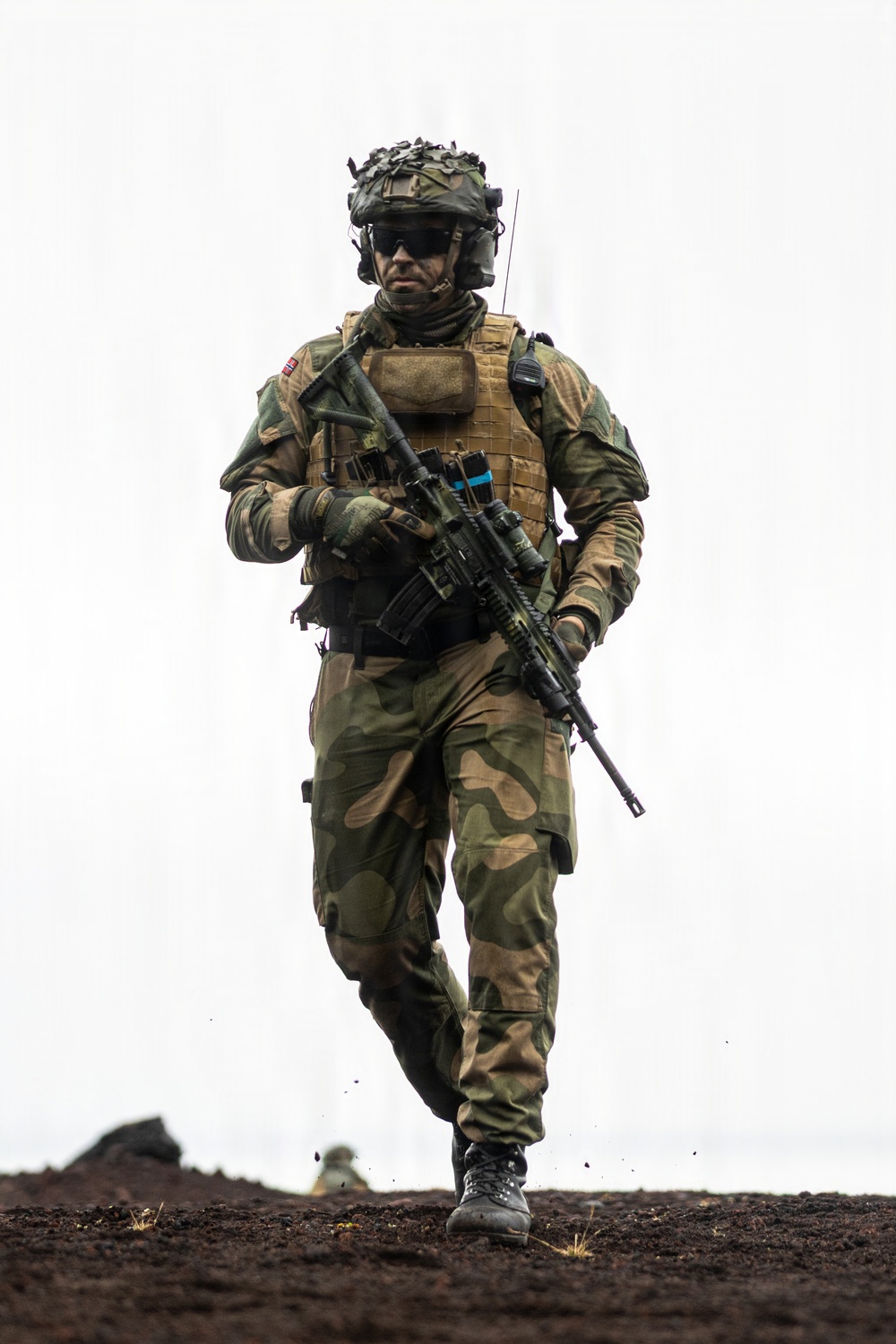 A fully equipped soldier in Norwegian M98 woodland camouflage uniform advances across dark, churned terrain. The soldier carries a scoped assault rifle and wears a plate carrier vest with a visible Norwegian flag patch on the left shoulder. The image is a high-quality professional military photograph taken from a low angle, giving the subject an imposing presence against an overcast sky.