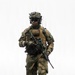 A fully equipped soldier in Norwegian M98 woodland camouflage uniform advances across dark, churned terrain. The soldier carries a scoped assault rifle and wears a plate carrier vest with a visible Norwegian flag patch on the left shoulder. The image is a high-quality professional military photograph taken from a low angle, giving the subject an imposing presence against an overcast sky.