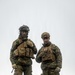 Two military personnel in full combat gear stand side by side on elevated terrain against an overcast sky. The left individual wears a woodland camouflage pattern uniform with a Norwegian flag patch visible on the shoulder, while the right individual wears MARPAT (Marine Pattern) digital camouflage consistent with U.S. Marine Corps uniforms. Both are equipped with tactical plate carriers, helmets with mounted accessories, and gloves, suggesting a joint multinational military exercise or operation.