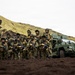A group of approximately 15 military personnel in camouflage uniforms pose for a unit photograph in a volcanic landscape. They are armed with assault rifles and positioned in front of a green armored military vehicle. The terrain features dark volcanic soil and moss-covered hills consistent with Icelandic or Nordic geography.