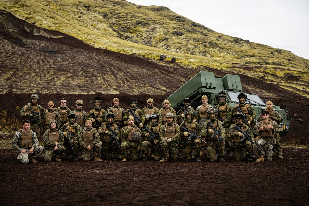 A group of approximately 25 military personnel in full combat gear pose for a group photograph in a volcanic landscape with dark soil and moss-covered hills. Behind them is a large green military vehicle, likely an M142 HIMARS or similar rocket artillery system. The setting and terrain suggest a northern or Arctic environment, possibly Iceland or a similar volcanic region.