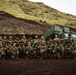 A group of approximately 25 military personnel in full combat gear pose for a group photograph in a volcanic landscape with dark soil and moss-covered hills. Behind them is a large green military vehicle, likely an M142 HIMARS or similar rocket artillery system. The setting and terrain suggest a northern or Arctic environment, possibly Iceland or a similar volcanic region.