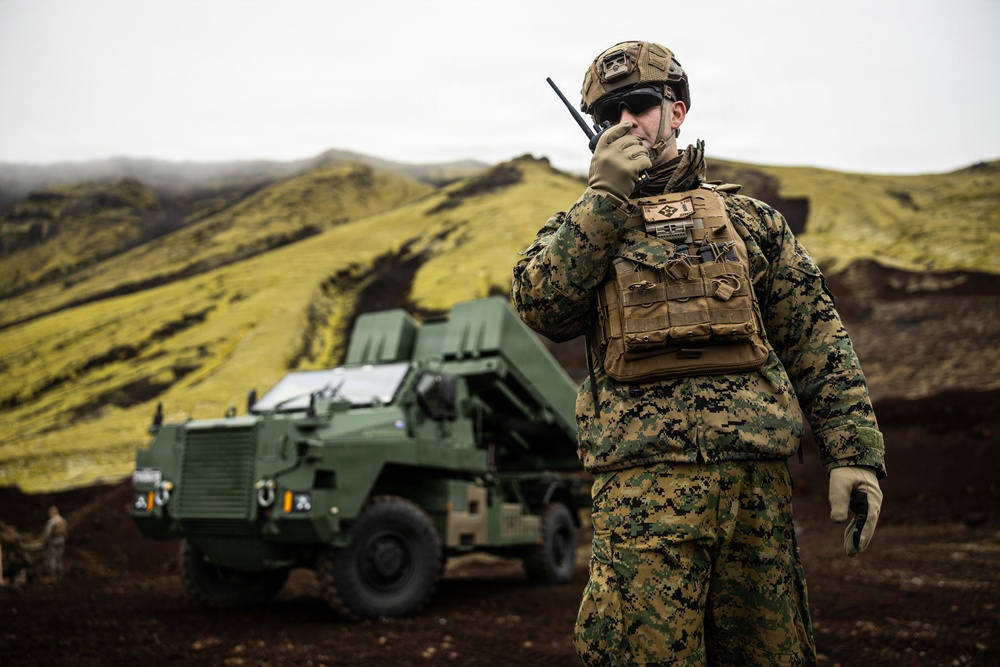 A military service member in digital camouflage uniform communicates via handheld radio in a rugged, volcanic terrain environment. Behind the soldier stands a large green military wheeled vehicle, likely a rocket artillery or missile launcher system. The landscape features dark volcanic soil and moss-covered green hills under an overcast sky.