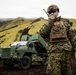 A military service member in digital camouflage uniform communicates via handheld radio in a rugged, volcanic terrain environment. Behind the soldier stands a large green military wheeled vehicle, likely a rocket artillery or missile launcher system. The landscape features dark volcanic soil and moss-covered green hills under an overcast sky.