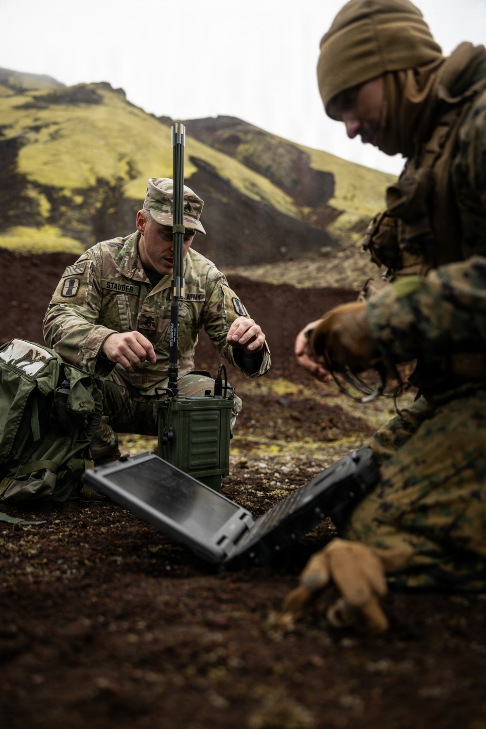 Two military personnel are setting up communications or electronic equipment in a rugged outdoor terrain with volcanic-looking hills in the background. The soldier on the left, identified by name tape as 'STAUDER' and wearing U.S. Army OCP (Operational Camouflage Pattern) uniform, is operating a green military radio/communications unit with antennas. A second individual in MARPAT (Marine pattern) camouflage and a tan beanie is working with a ruggedized military laptop.