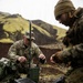 Two military personnel are setting up communications or electronic equipment in a rugged outdoor terrain with volcanic-looking hills in the background. The soldier on the left, identified by name tape as 'STAUDER' and wearing U.S. Army OCP (Operational Camouflage Pattern) uniform, is operating a green military radio/communications unit with antennas. A second individual in MARPAT (Marine pattern) camouflage and a tan beanie is working with a ruggedized military laptop.