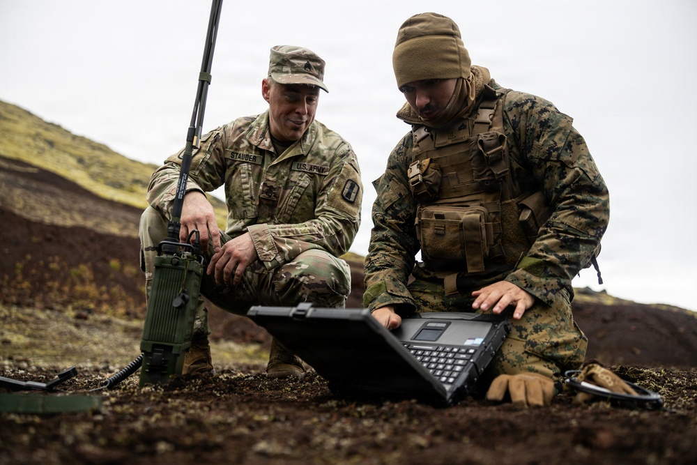 Two military personnel from different branches are working together in a field environment, examining a ruggedized laptop computer. The soldier on the left wears U.S. Army OCP (Operational Camouflage Pattern) uniform with a name tape reading 'STAUDER,' while the individual on the right wears USMC MARPAT digital camouflage with tactical vest. They appear to be operating communications or command equipment in a rocky, volcanic-looking outdoor terrain.