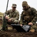 Two military personnel from different branches are working together in a field environment, examining a ruggedized laptop computer. The soldier on the left wears U.S. Army OCP (Operational Camouflage Pattern) uniform with a name tape reading 'STAUDER,' while the individual on the right wears USMC MARPAT digital camouflage with tactical vest. They appear to be operating communications or command equipment in a rocky, volcanic-looking outdoor terrain.