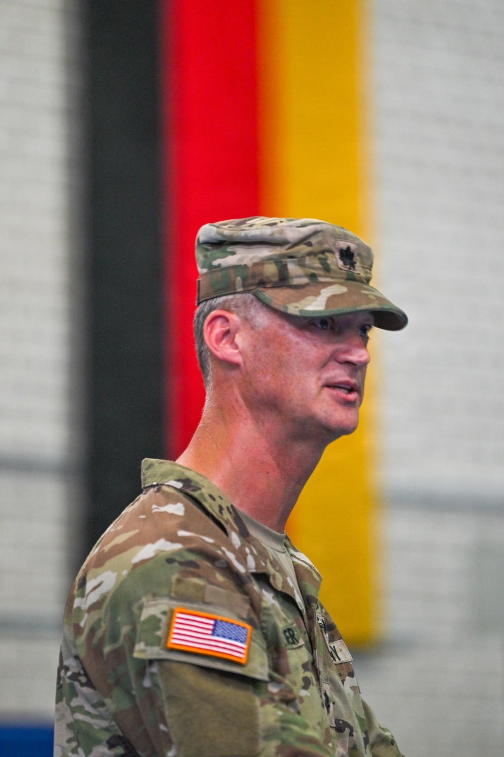 A U.S. Army officer in MultiCam (OCP) uniform is photographed in profile, appearing to be speaking at a formal military event. A German national flag (black, red, and gold stripes) is visible in the blurred background, suggesting the event is taking place in Germany, likely at a U.S. military installation. The officer wears a rank insignia on his cap consistent with a Major (oak leaf cluster).