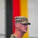 A U.S. Army officer in MultiCam (OCP) uniform is photographed in profile, appearing to be speaking at a formal military event. A German national flag (black, red, and gold stripes) is visible in the blurred background, suggesting the event is taking place in Germany, likely at a U.S. military installation. The officer wears a rank insignia on his cap consistent with a Major (oak leaf cluster).