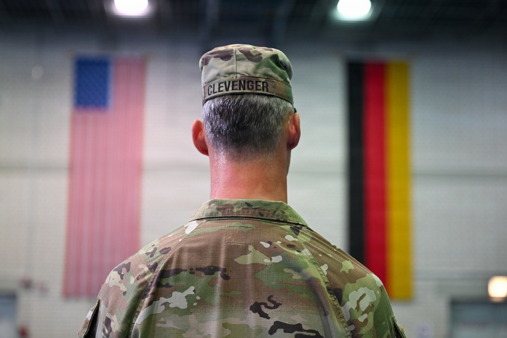 A U.S. military soldier named Clevenger is photographed from behind, standing at attention in what appears to be a large indoor facility such as a gymnasium or hangar. Two flags are visible in the background — the United States flag on the left and the German flag on the right — suggesting a joint military or bilateral event. The soldier is wearing OCP (Operational Camouflage Pattern) uniform with a patrol cap bearing the name tape 'CLEVENGER'.