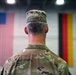A U.S. military soldier named Clevenger is photographed from behind, standing at attention in what appears to be a large indoor facility such as a gymnasium or hangar. Two flags are visible in the background — the United States flag on the left and the German flag on the right — suggesting a joint military or bilateral event. The soldier is wearing OCP (Operational Camouflage Pattern) uniform with a patrol cap bearing the name tape 'CLEVENGER'.