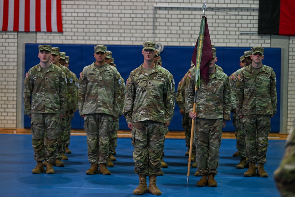 A formation of U.S. Army soldiers in Operational Camouflage Pattern (OCP) uniforms stands at attention inside what appears to be a gymnasium or indoor drill hall. A soldier in the foreground center holds a guidon (unit flag) with a maroon/burgundy pennant on a wooden staff. Two flags are visible on the wall behind them, including the American flag and what appears to be the Afghan national flag, suggesting this may be a deployment or redeployment ceremony.
