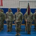A formation of U.S. Army soldiers in Operational Camouflage Pattern (OCP) uniforms stands at attention inside what appears to be a gymnasium or indoor drill hall. A soldier in the foreground center holds a guidon (unit flag) with a maroon/burgundy pennant on a wooden staff. Two flags are visible on the wall behind them, including the American flag and what appears to be the Afghan national flag, suggesting this may be a deployment or redeployment ceremony.