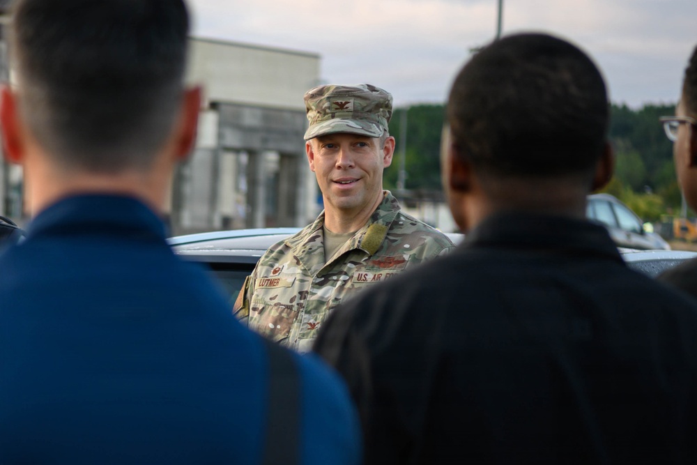 A U.S. Air Force officer in OCP (Operational Camouflage Pattern) uniform is addressing a small group of individuals outdoors. The officer's name tape reads 'LUTMER' and the branch tape reads 'U.S. AIR FORCE,' with a Colonel's eagle insignia visible on the patrol cap. The setting appears to be a military installation or base, with buildings and vehicles visible in the background.