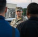 A U.S. Air Force officer in OCP (Operational Camouflage Pattern) uniform is addressing a small group of individuals outdoors. The officer's name tape reads 'LUTMER' and the branch tape reads 'U.S. AIR FORCE,' with a Colonel's eagle insignia visible on the patrol cap. The setting appears to be a military installation or base, with buildings and vehicles visible in the background.