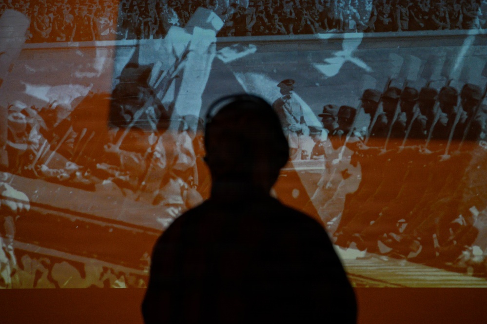 A silhouetted figure stands in front of a large projected archival image showing what appears to be a military parade or march. The projection is tinted in orange and blue tones, displaying uniformed soldiers carrying rifles or bayonets. The composition creates a dramatic contrast between the dark foreground silhouette and the historical imagery behind.