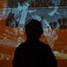 A silhouetted figure stands in front of a large projected archival image showing what appears to be a military parade or march. The projection is tinted in orange and blue tones, displaying uniformed soldiers carrying rifles or bayonets. The composition creates a dramatic contrast between the dark foreground silhouette and the historical imagery behind.