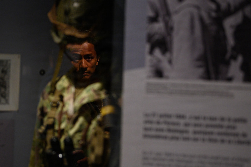 A museum display featuring a military mannequin dressed in camouflage combat gear, positioned behind glass in a dimly lit exhibit case. To the right, an informational panel with text and a black-and-white archival photograph is visible, though both are partially out of focus. The exhibit appears to document military history, likely related to a specific conflict or soldier's experience.